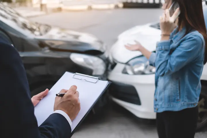 Person filling in a form on a clipboard while a woman on the phone stands by two damaged cars